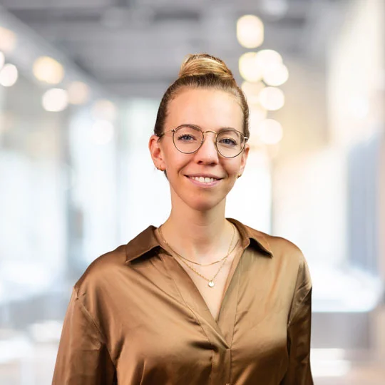 Porträt von Nicole Schröder, Projektmanagerin bei ITS mobility, in einer modernen, professionellen Umgebung. Sie trägt eine Brille und ein elegantes, beiges Hemd, mit unscharfem Hintergrund und warmen Lichtern, die eine freundliche Atmosphäre schaffen